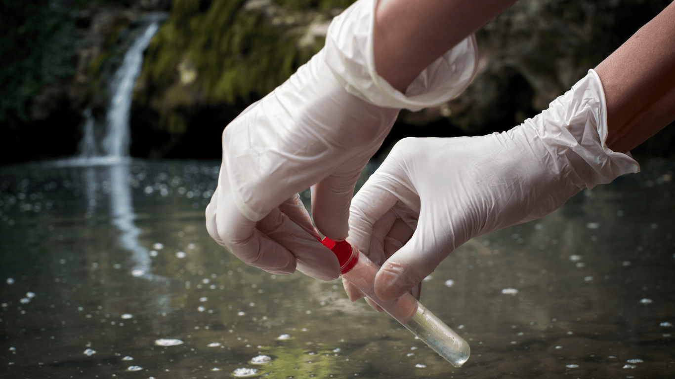 Gloved hands collecting a water sample from a natural water source for water treatment chemicals analysis, illustrating professional water treatment and wastewater treatment processes used by industrial business services and supply providers to support customers and water systems. The image represents how treatment chemicals such as ferric chloride, ferric sulfate, hydrogen peroxide, chlorine dioxide, chlorine, calcium hydroxide, phosphoric acid, soda ash, salts, acids, iron, and sulfur are utilized in treating water and wastewater to achieve pH adjustment, bacteria control, and contamination removal. This sampling process supports safe drinking water production, wastewater compliance, and industrial treatment applications, including paper manufacturing, where treatment chemicals are used as a precursor to effective disinfection, oxidation, coagulation, and system protection. The scene reflects how water treatment services utilize chemical treatment solutions to manage wastewater, control bacteria, adjust pH, and ensure reliable water treatment for industrial customers and businesses focused on safe, compliant, and efficient water systems.